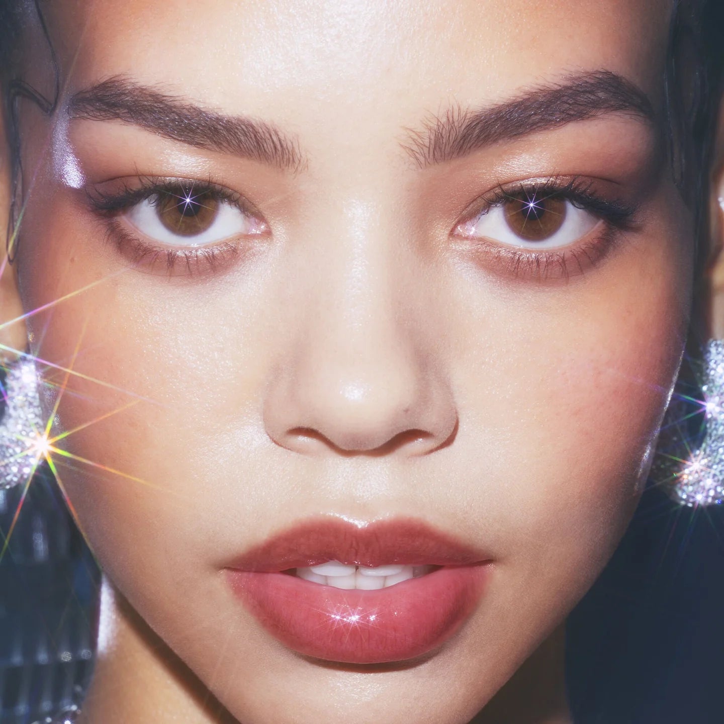 Close-up portrait of a woman with defined eyebrows, brown eyeshadow, glossy pinkish-red lips, and sparkling silver earrings against a dark background with starburst light effects.